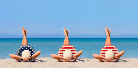 Three young girls on the beach wearing straw hats in the colors of the flag of USA. The concept of the perfect holiday in the resorts of the United States. Focus on hats.の写真素材