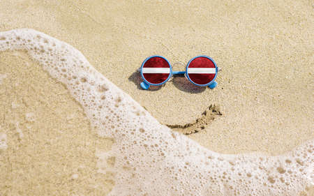 Sunglasses with flag of Latvia on a sandy beach. Nearby is a sea lightning and a painted smile. The concept of a successful vacation in the resorts of Latvia.の写真素材