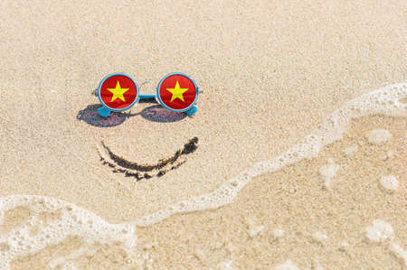 A painted smile on the sand and sunglasses with the flag of the Vietnam. The concept of a positive and successful holiday in the resort of Vietnam.の写真素材