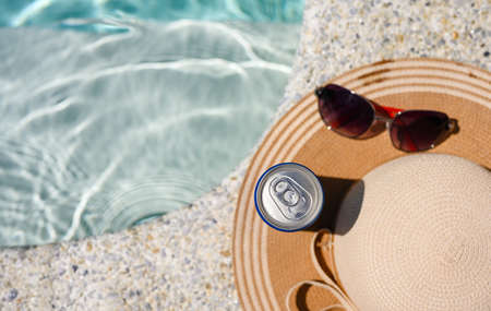 A can of cold drink stands by the hotel pool next to a straw hat and sunglasses. Top view, selective focus on a can of drink.の写真素材