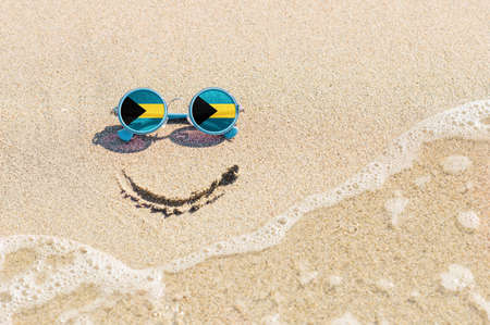 A painted smile on the beach and sunglasses with the flag of the Bahamas. The concept of a positive holiday in the resort of the Bahamas.の写真素材