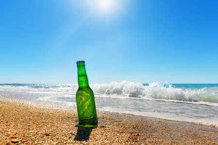 A bottle of beer on the beach against the backdrop of sea waves and bright sun.の写真素材