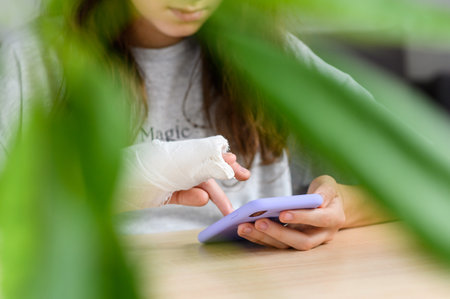 A teenage girl with a broken arm with a smartphone in her hands. Close-up of a girl with a bandage on her arm uses the phone. Selective focus on the fingers and bandage.の写真素材
