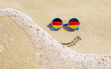 Sunglasses with flag of Germany on a sandy beach. Nearby is a sea lightning and a painted smile. Travel and vacation concept for Germansの写真素材