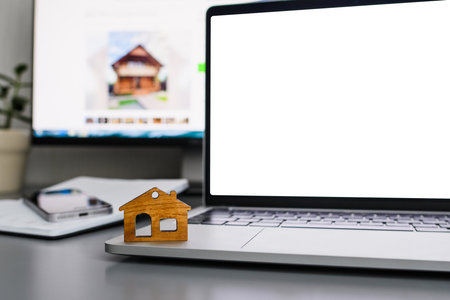 The concept of a service for the selection or rental of housing. Wooden figurine of a house on the background of a laptop with a white displayの写真素材