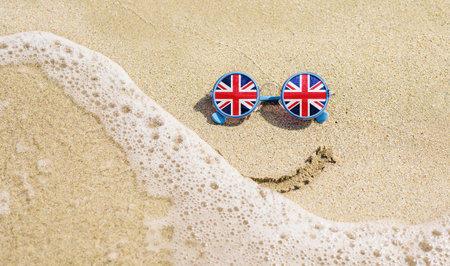 Sunglasses with flag of Great Britain on a sandy beach. Nearby is a sea lightning and a painted smile. Travel and vacation concept for Britonsの写真素材
