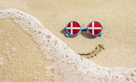 Sunglasses with flag of Denmark on a sandy beach. Nearby is a sea lightning and a painted smile. travel and vacation conceptの写真素材