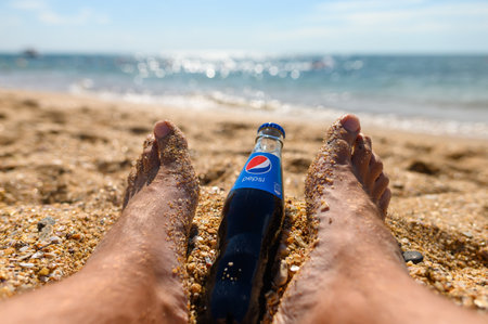 Anapa - Russia, July 2022: A bottle of Pepsi Cola lies near the male feet against the backdrop of the beach and the sea. Selective focus on the logo. Relax concept.のeditorial素材