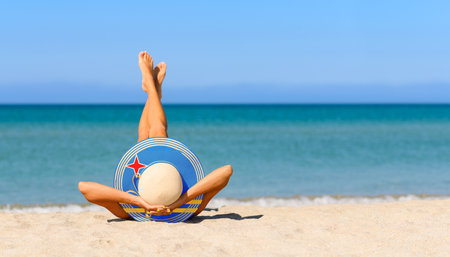 A slender girl on the beach in a straw hat in the colors of the Aruba flag. The concept of a perfect vacation in a resort in the Aruba. Focus on the hat.の写真素材
