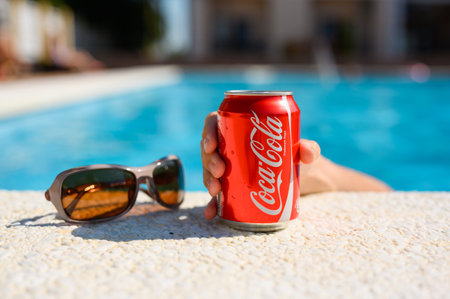 Anapa, Russia - July 18, 2023: The hand of a vacationing tourist reaches for a can of Coca Cola by the pool. Hot summer day by the pool. Close-up, selective focusのeditorial素材