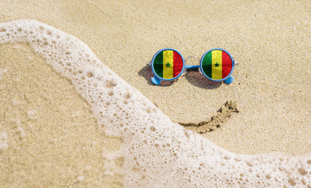 Sunglasses with flag of Senegal on a sandy beach. Nearby is a sea lightning and a painted smile. The concept of a successful vacation in the resorts of the Senegal.の写真素材