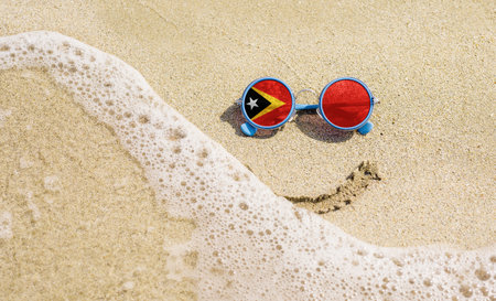 Sunglasses with flag of East Timor on a sandy beach. Nearby is a sea lightning and a painted smile.の写真素材