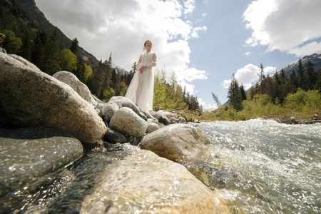 Happy bride in a white dress, a wreath of live cubs and leaves and a bunch of flowers on the shore of a mountain riverの写真素材