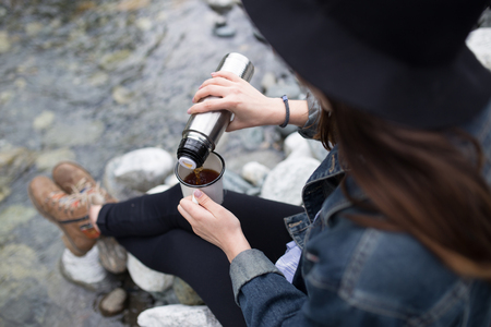 Girl wearing backpack drinking tea from flask and looking at the lakeの写真素材