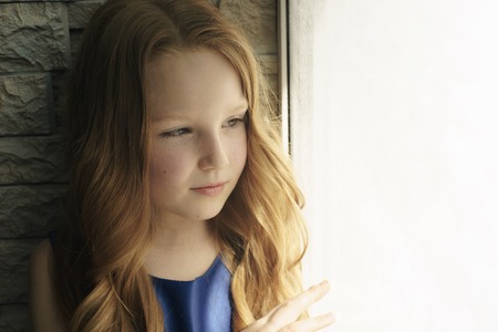 Hopeless life. Close up of depressed poor little girl standing near window and looking aside while feeling miserableの写真素材