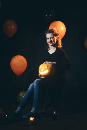 Halloween Witch with a carved Pumpkin and magic lights in a dark forest. Beautiful young surprised woman in witches hat and costume holding pumpkin. Halloween party artの写真素材