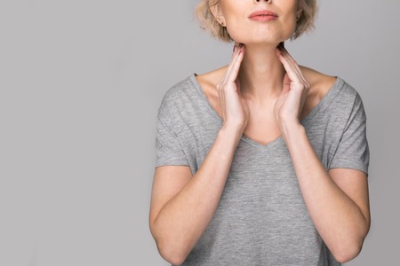 Female checking thyroid gland by herself. Close up of woman in white t- shirt touching neck with red spot. Thyroid disorder includes goiter, hyperthyroid, hypothyroid, tumor or cancer. Health care.の写真素材