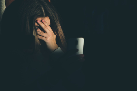 Single sad teen holding a mobile phone lamenting sitting on the bed in her bedroom with a dark lightの写真素材