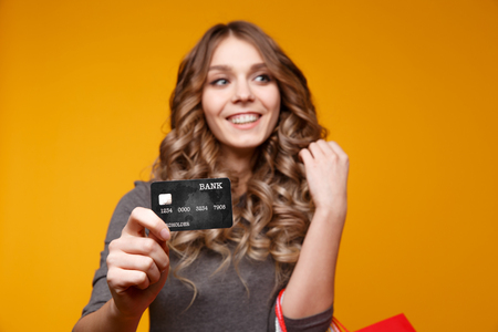 Portrait of a surprised happy girl holding shopping bags and showing credit card while looking at camera isolated over yellowの写真素材