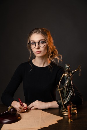 Young lawyer woman in a formal dress and spectacles sitting at table and writing something by pen,...