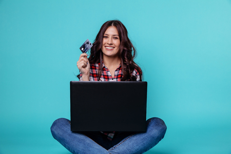 Smiling brunette woman sitting on the flour with laptop and credit card to make online shopping.の写真素材