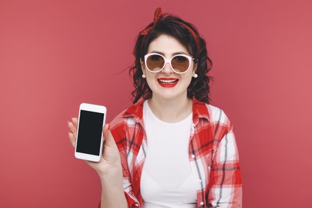 Portrait of an excited beautiful girl wearing red shirt, holding mobile phone isolated over pink...