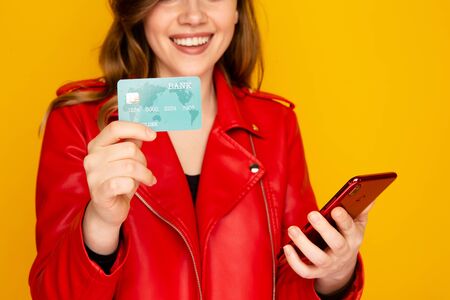 Woman in the red jacket holding credit card and phone isolated over the yellow background.