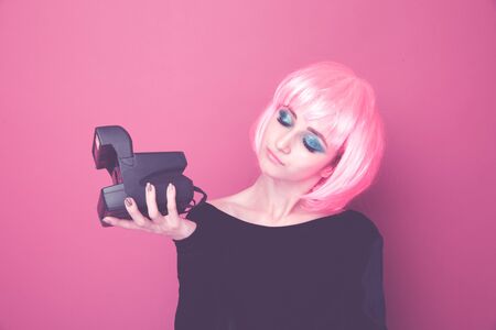 Young stylish woman holding retro camera and standing in a pink wig in a bright studio.の写真素材