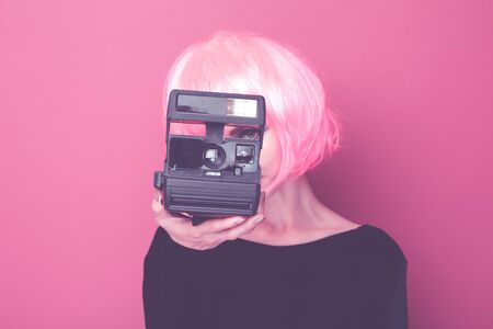 Young stylish woman holding retro camera and standing in a pink wig in a bright studio.