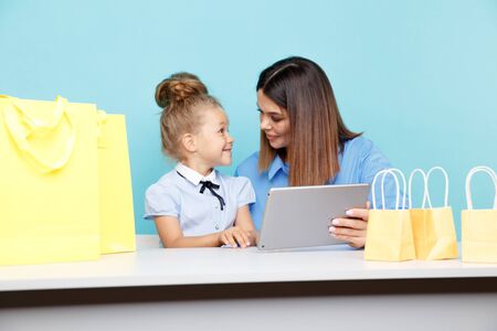 Portrait of mother and daughter with tablet sitting at the table isolated over the blue studio. Online family shopping concept.の写真素材