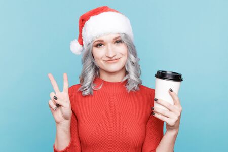Cheerful female model in the red santa claus hat holding a cup of coffee and smiling in the studio.の写真素材