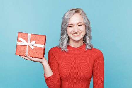 Exited smiling young woman in red dress holding gift box in the blue studio.の写真素材