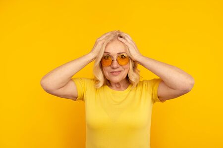 Exited pretty adult female in yellow T-shut and orange sunglasses with her hands on head.