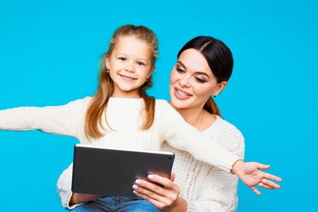 Portrait of mother and daughter with tablet sitting on the floor isolated over the blue studio.