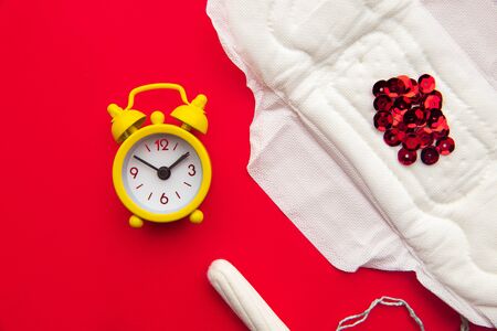 Close-up of feminine pad and cotton tampon with red glitters and yellow alarm clock on red backgroundの写真素材