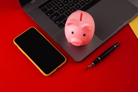 Moneybox near laptop and smartphone, place for text. Finance and budget concept. Piggy bank in pink color with gadgets and stationery on colorful backgroundの写真素材