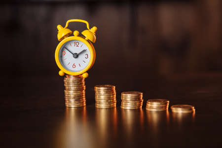 Time is money concept. An alarm clock and stack of coins on a wooden tableの写真素材