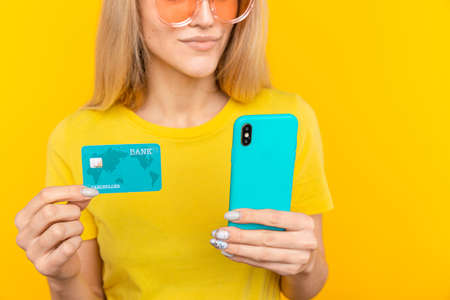 Portrait of a young blonde girl showing plastic credit card while using mobile phone isolated over...