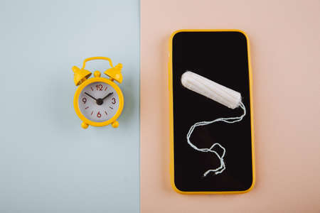 Menstrual cycle concept. Yellow alarm clock and smartphone screen with cotton tamponの写真素材