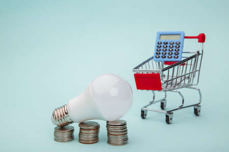 Light bulb with coins stack and trolley with calculator on blue background. Energy saving conceptの写真素材
