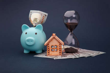 Blue piggy bank with wooden model of house, hourglass and money banknotes on blue background. Savings money for buy houseの写真素材
