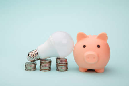 Piggy bank, stack of coins and led light lamp on blue background. Energy saving conceptの写真素材