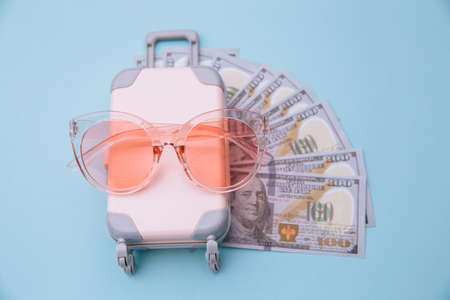 Mini travel luggage suitcase, sunglasses and money banknotes on blue background. Savings for traveling conceptの写真素材