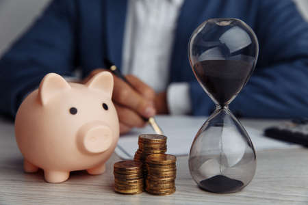 Businessman approves important contract in office. Pink piggy bank with stack of coins and hourglass on desk. Time is moneyの写真素材