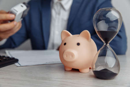 Businessman approves contract in office. Piggy bank with stack of coins and hourglass on desk close-up. Time is money conceptの写真素材