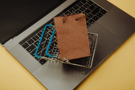 Concept of online shopping. Paper shopping bag and cart on keyboard of laptop isolated on yellow. Top viewの写真素材