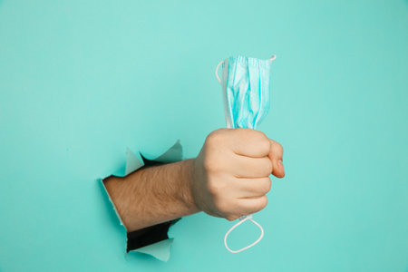 A male hand holds a medical mask through a hole in a blue wall. Medical breakthrough conceptの写真素材