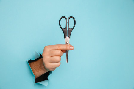 Scissors in male hand from a torn hole in blue backgroundの写真素材