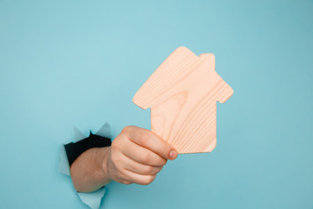 Mans hand with model of house through a hole in blue paper. House sale and rent conceptの写真素材