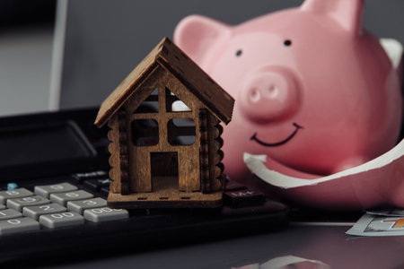 Concept of money savings for a new house. Broken piggy bank with calculator and wooden houseの写真素材